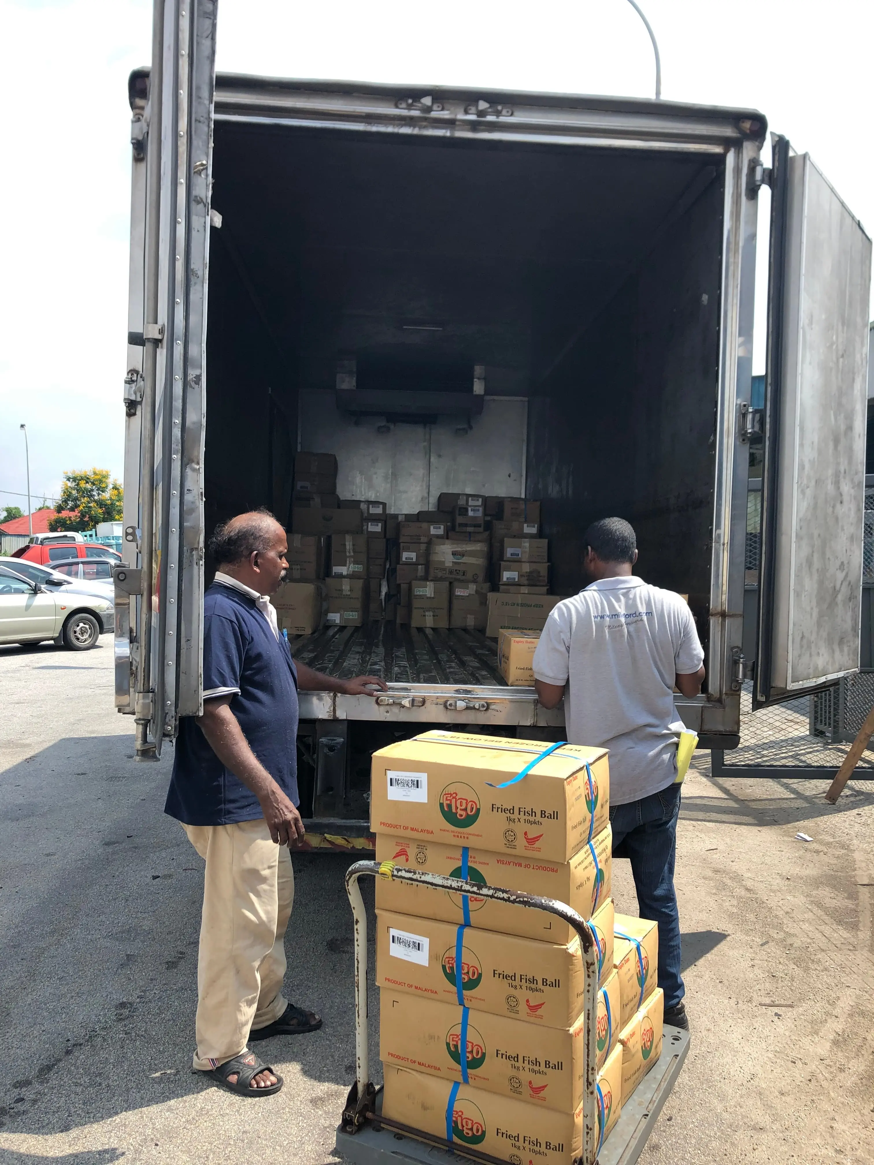 Mileford staff loading boxes of 'Fried Fish Ball' using a hand truck into a lorry, demonstrating careful handling.